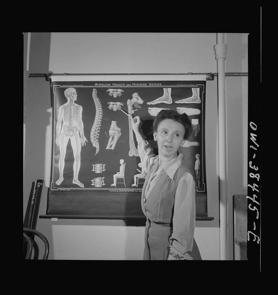 Black and white photo of a woman standing in front of an anatomy poster. She is posed as though she is teaching from the poster. 